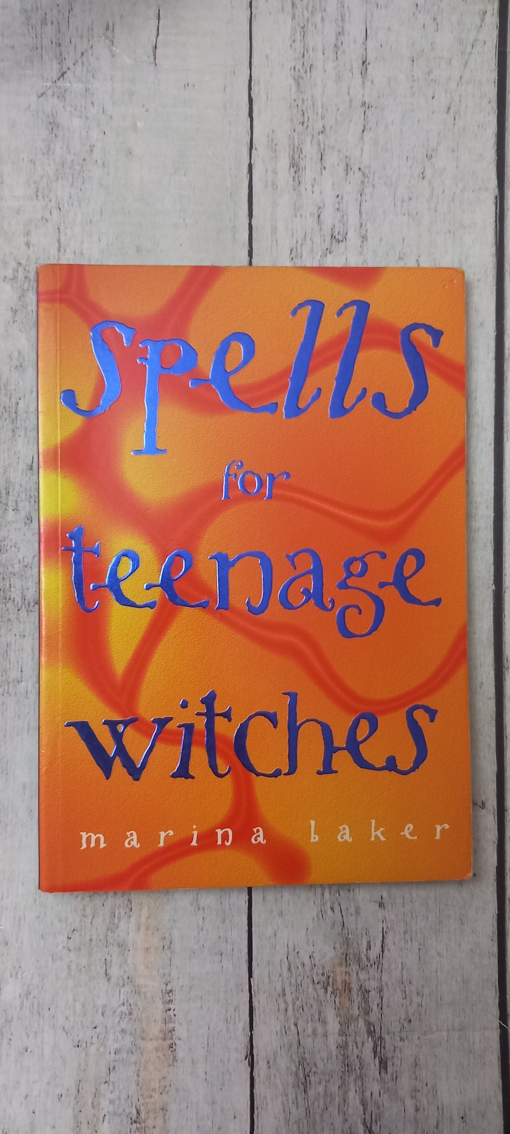 A book titled 'Spells for Teenage Witches' by Marina Laker, featuring a vibrant orange and yellow abstract patterned cover with blue and white text, placed on a light gray wooden surface.