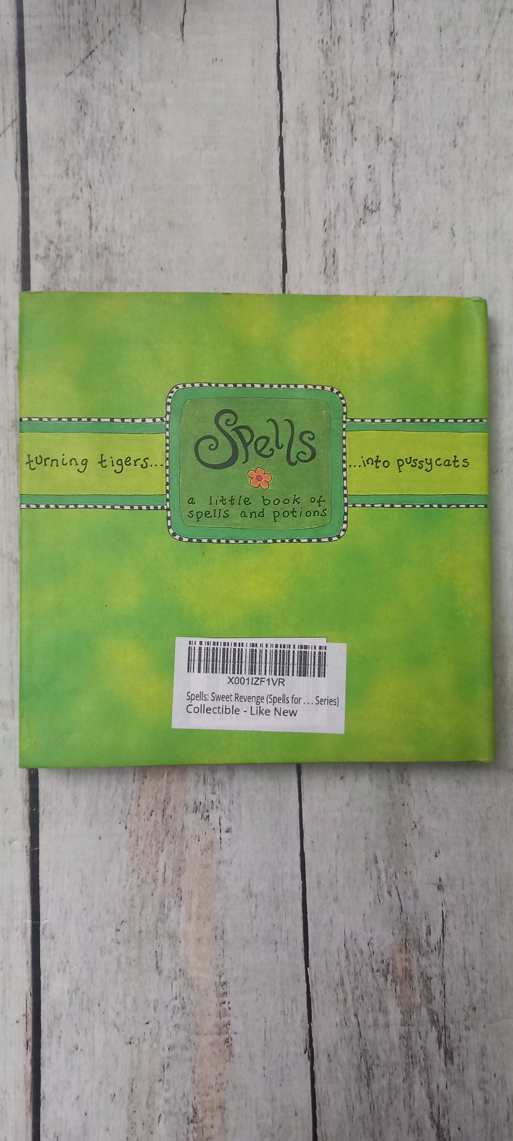 A square green card or paper product with a stitched-edge border and a central label reading 'Spells' and 'The Enchanted', photographed on a light wood surface with a barcode sticker on the right side.