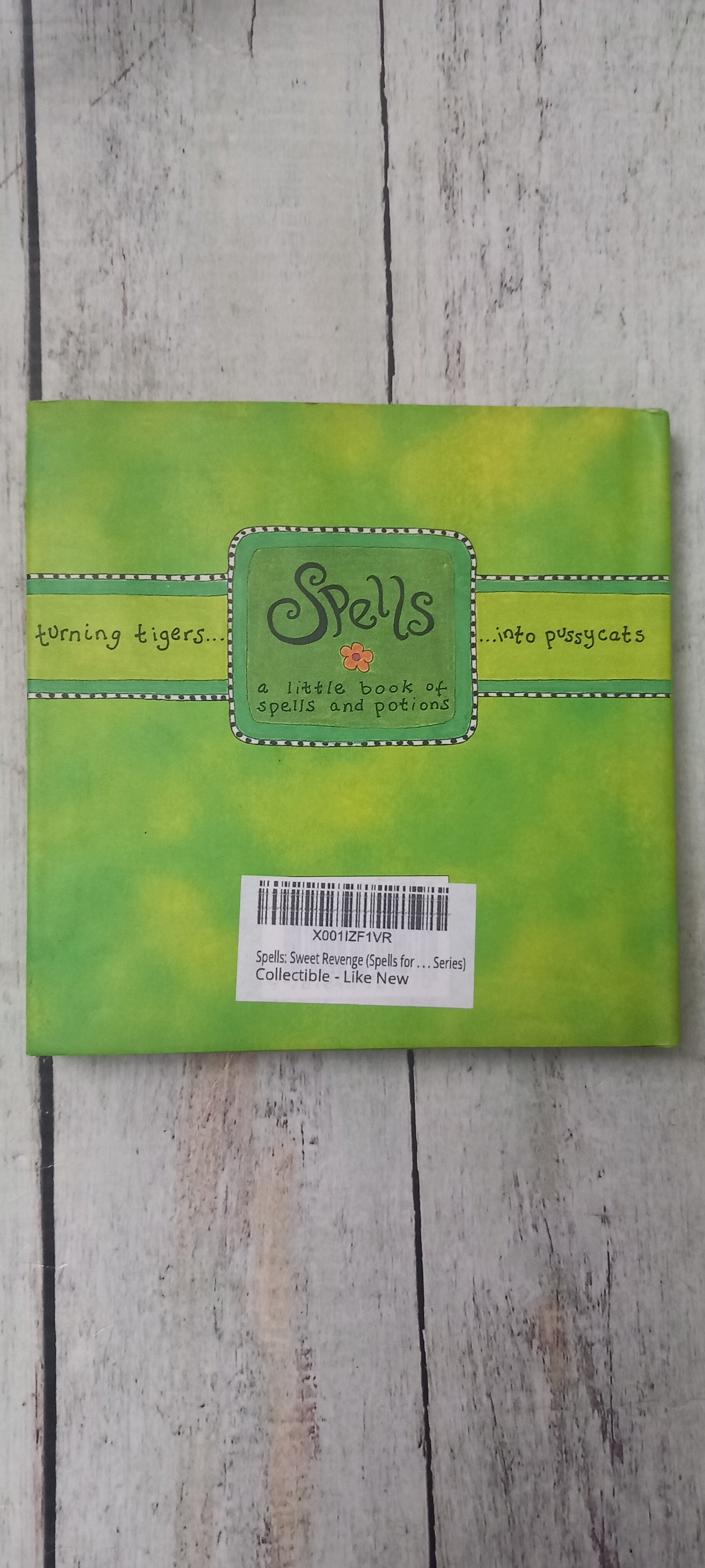 A square green card or paper product with a stitched-edge border and a central label reading 'Spells' and 'The Enchanted', photographed on a light wood surface with a barcode sticker on the right side.