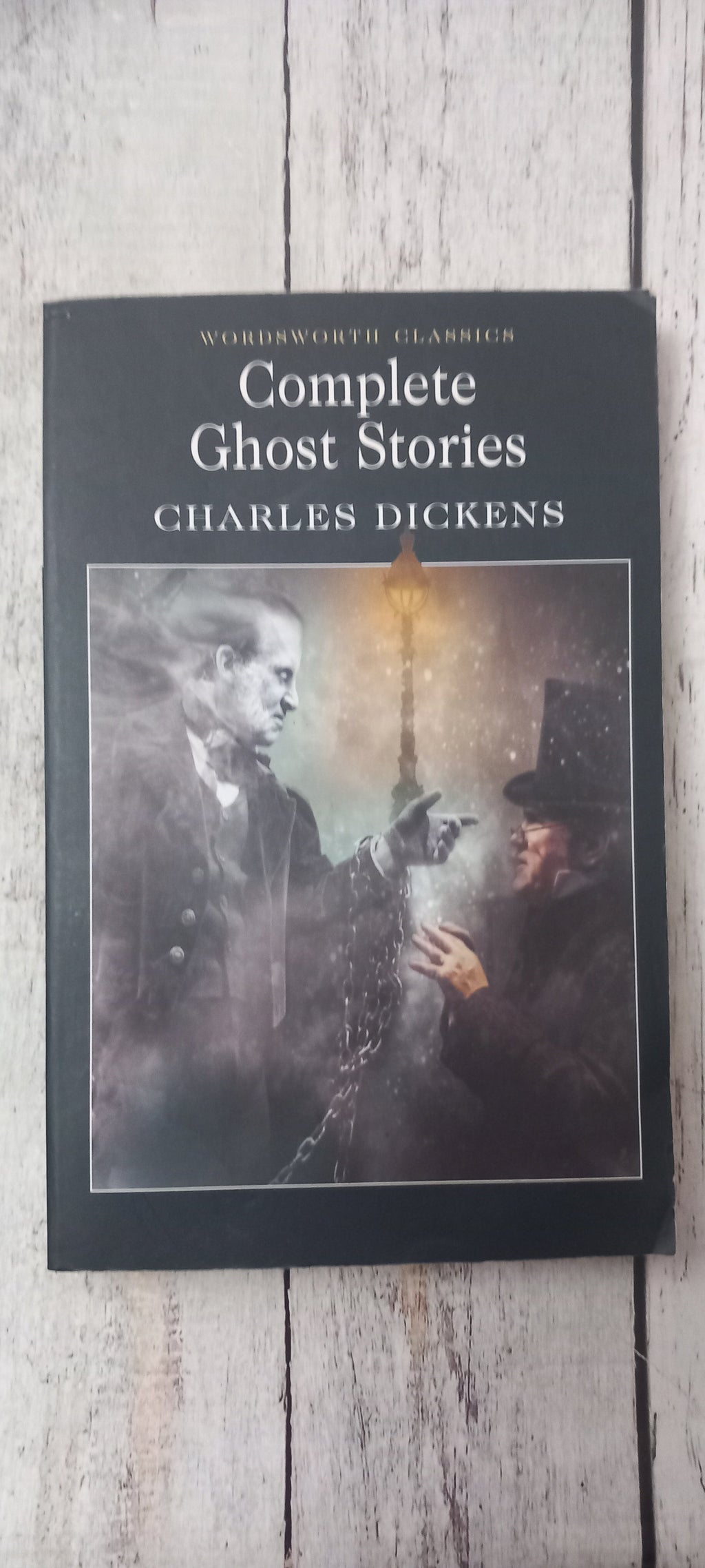 The book cover of 'Complete Ghost Stories' by Charles Dickens, featuring a dark, moody illustration of a ghostly figure and a lantern, displayed on a light-colored wooden surface.