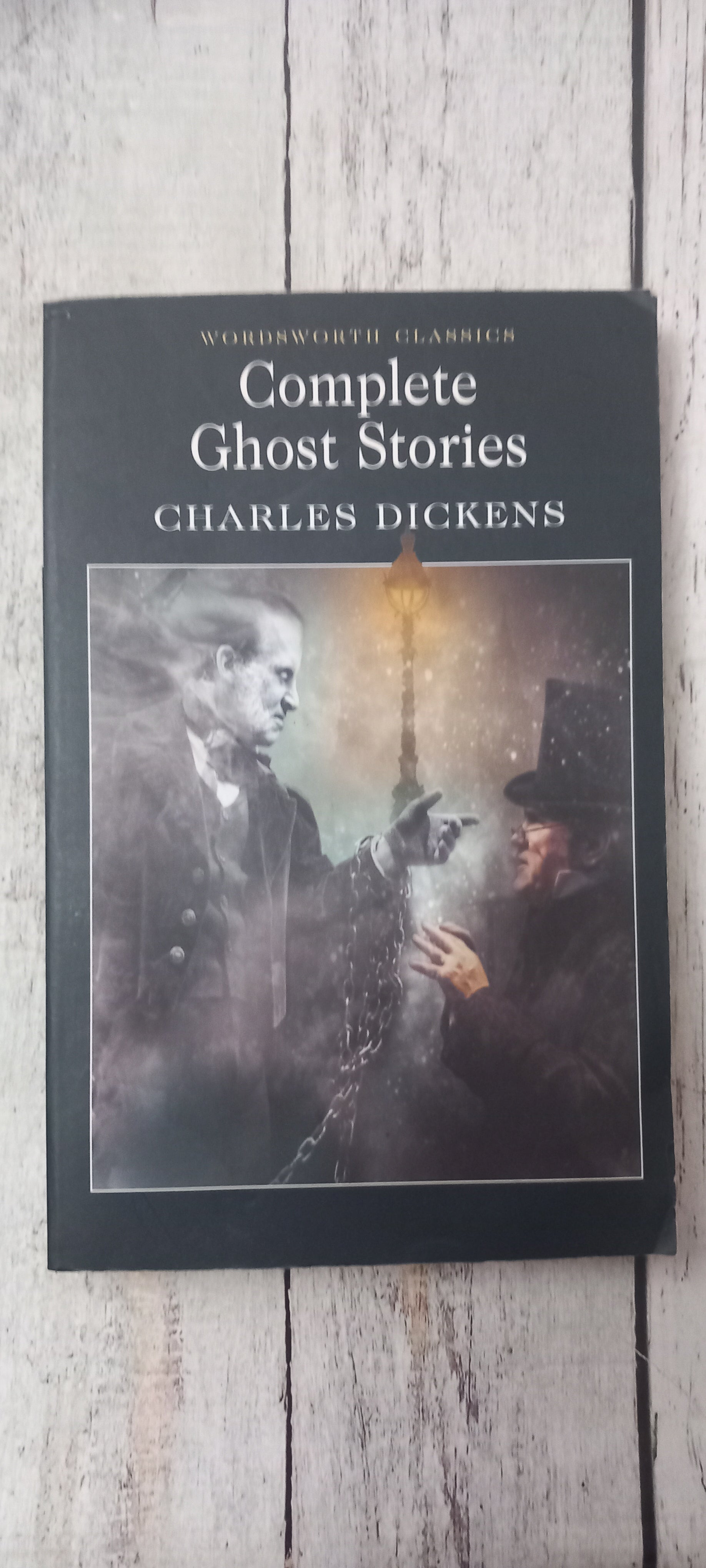 The book cover of 'Complete Ghost Stories' by Charles Dickens, featuring a dark, moody illustration of a ghostly figure and a lantern, displayed on a light-colored wooden surface.