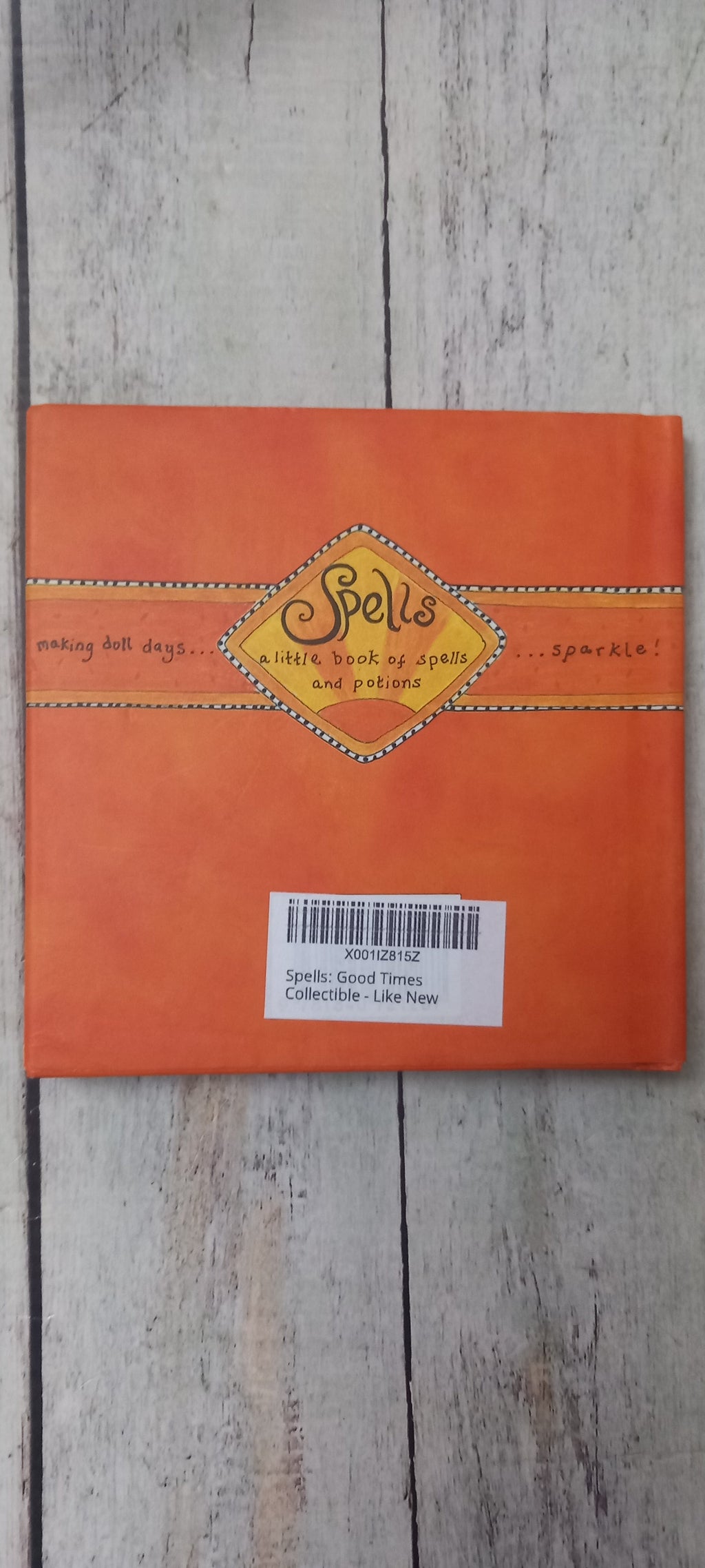 An orange square book or booklet titled 'Spells' with a yellow diamond-shaped label, photographed from above on a light weathered wood surface. A white barcode sticker is attached to the right side.
