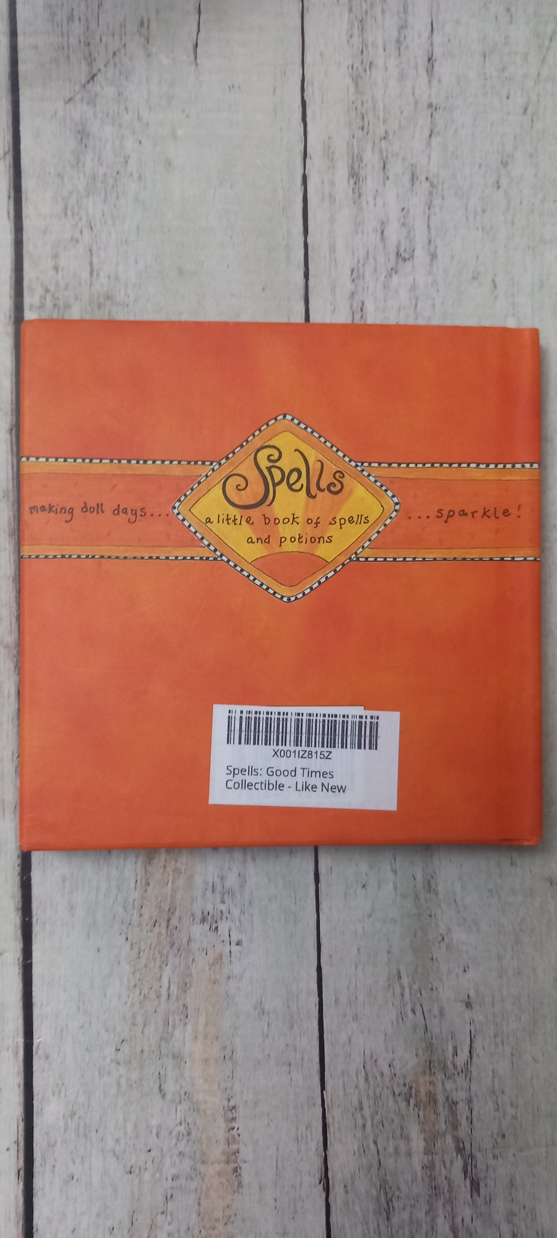 An orange square book or booklet titled 'Spells' with a yellow diamond-shaped label, photographed from above on a light weathered wood surface. A white barcode sticker is attached to the right side.