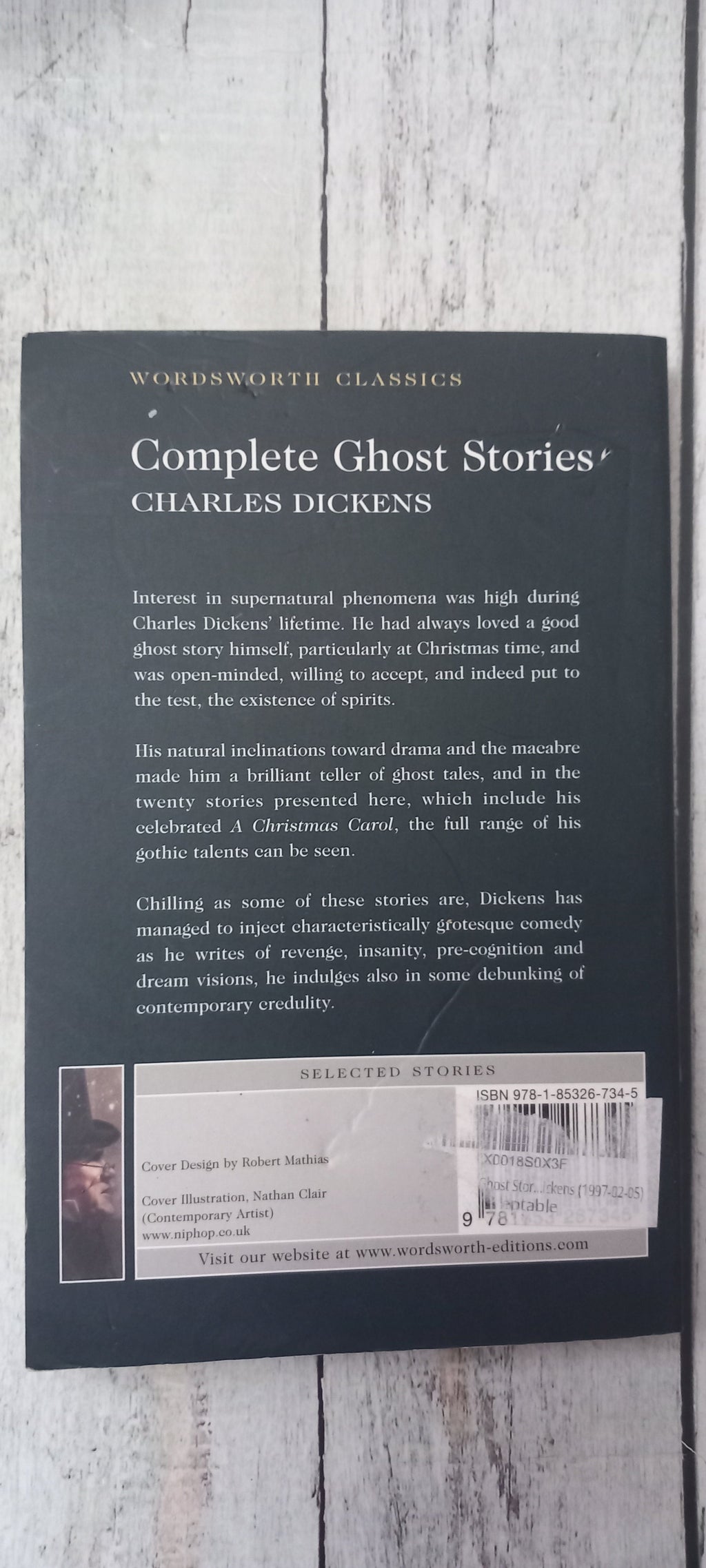 The back cover of the book 'Complete Ghost Stories' by Charles Dickens, featuring a black background with white text and a barcode, photographed on a light gray wood plank surface.