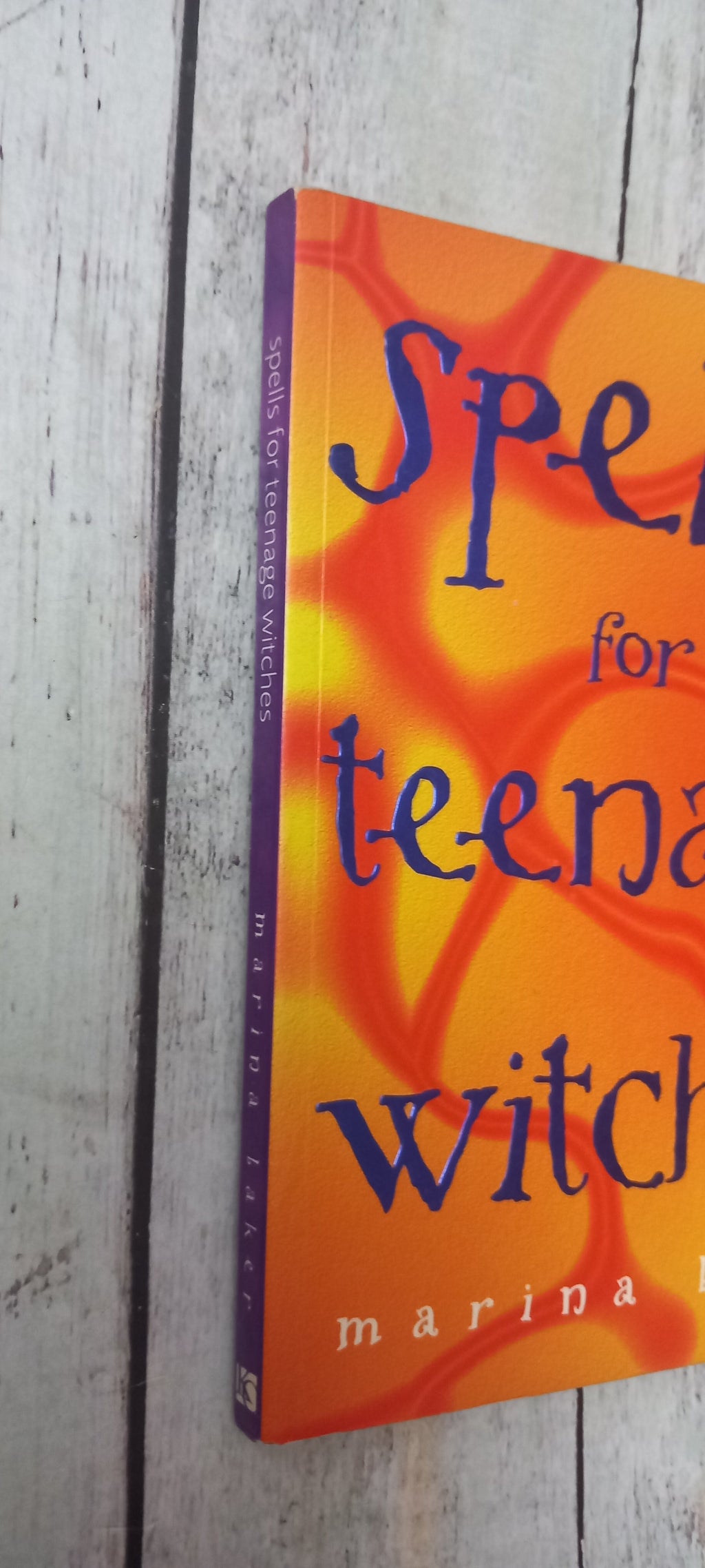 A book titled 'Spells for Teenage Witches' by Marina Warner, featuring a vibrant orange and yellow cover with dark blue text, photographed on a light gray wooden surface.