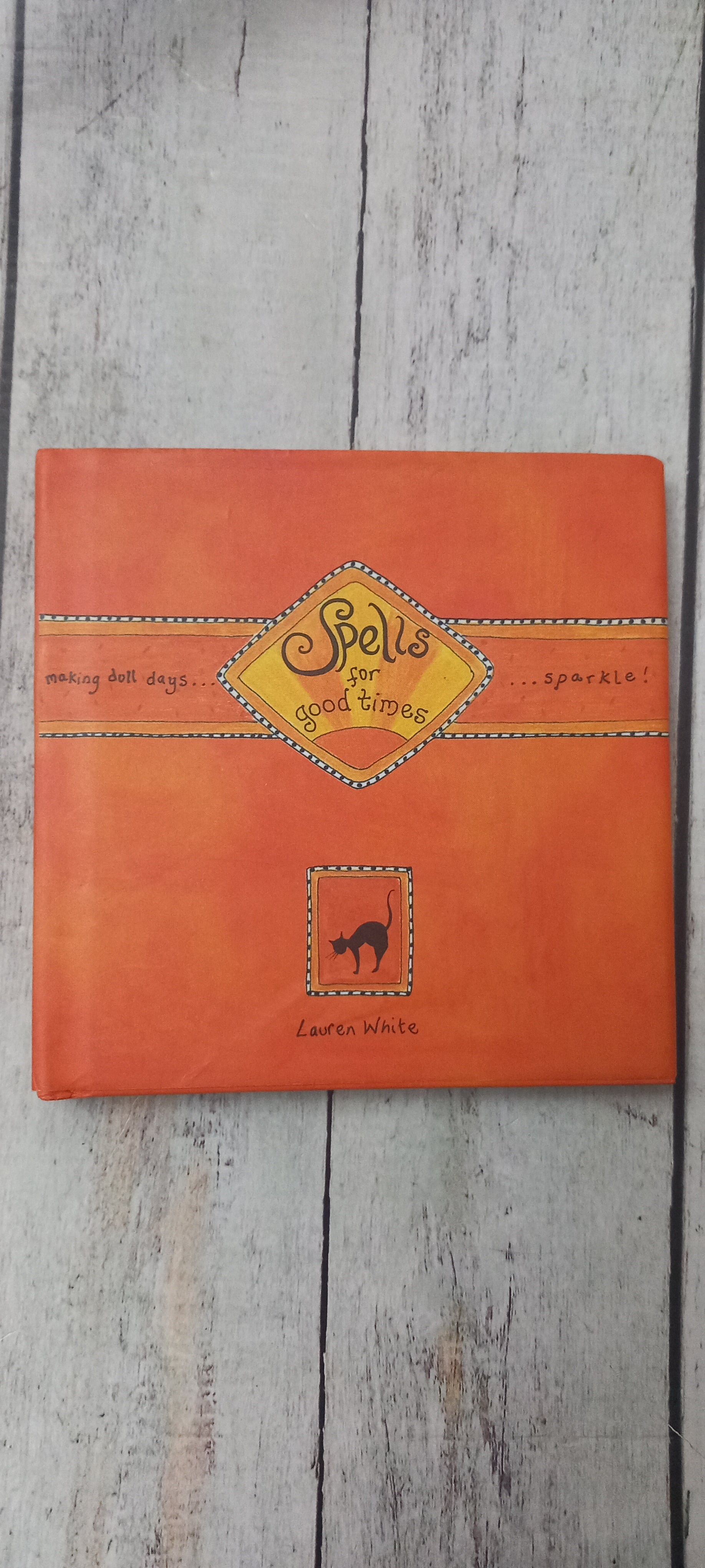 An orange hardcover book titled 'Bells for Bad Boys' by Laura White, featuring a yellow diamond-shaped title label and a small black cat silhouette, displayed on a light gray wood plank surface.